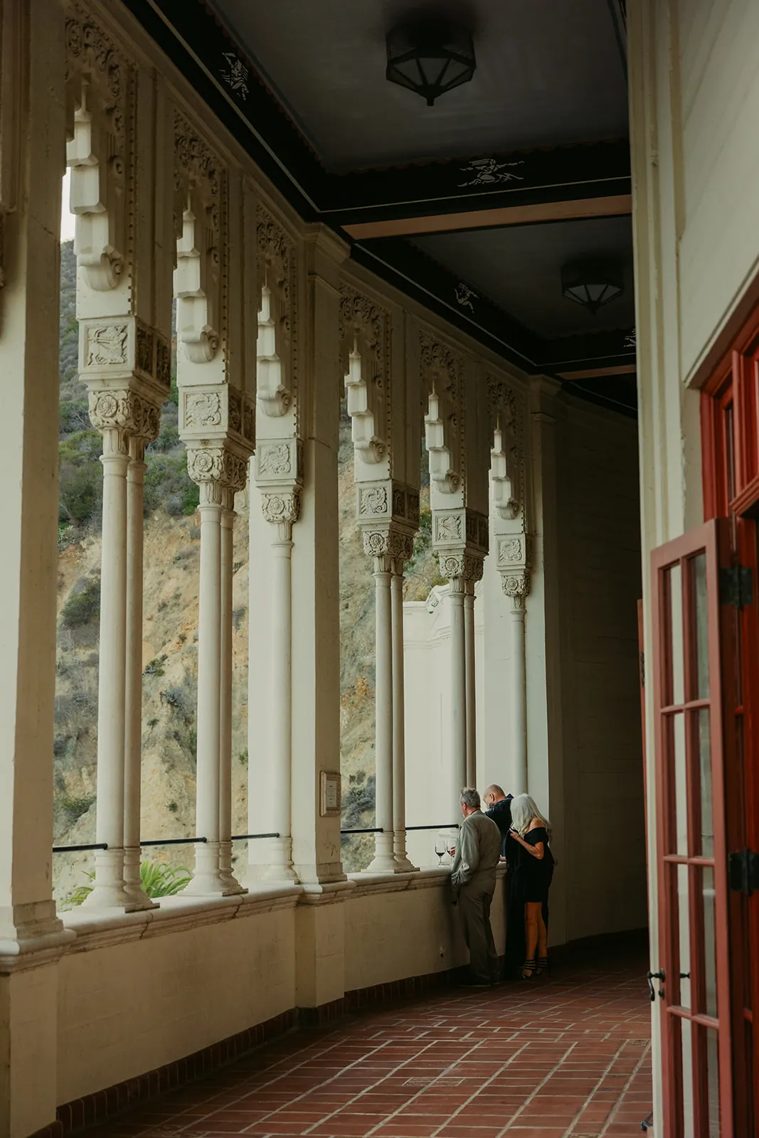 Couple standing in the ornate Casino colonnade corridor during cocktail hour at their Catalina Island Casino wedding, white arched columns and red doors overlooking the hillside
