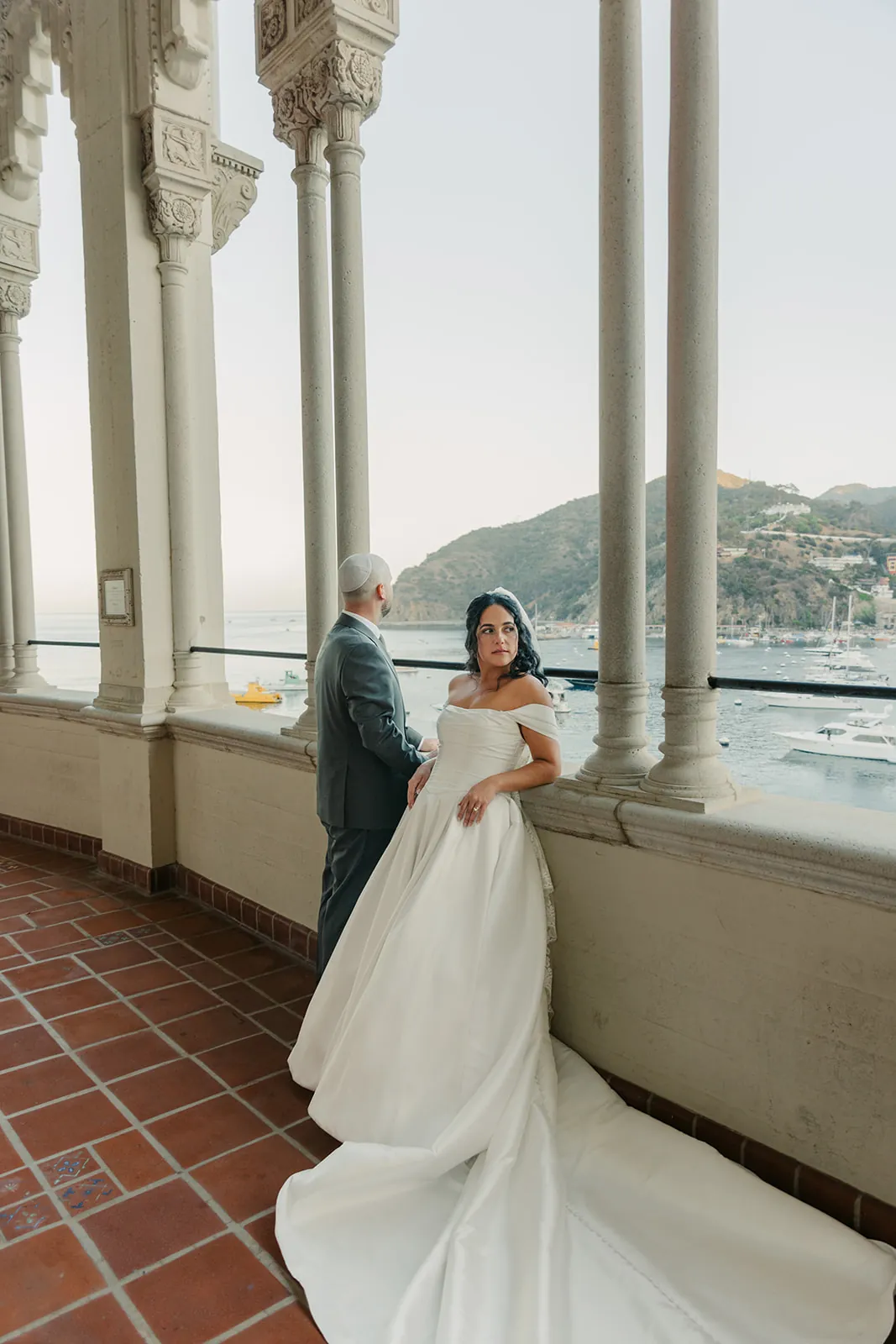 Bride leaning against the Casino colonnade railing with groom behind her, Avalon Bay and Catalina hills in the background at their destination wedding