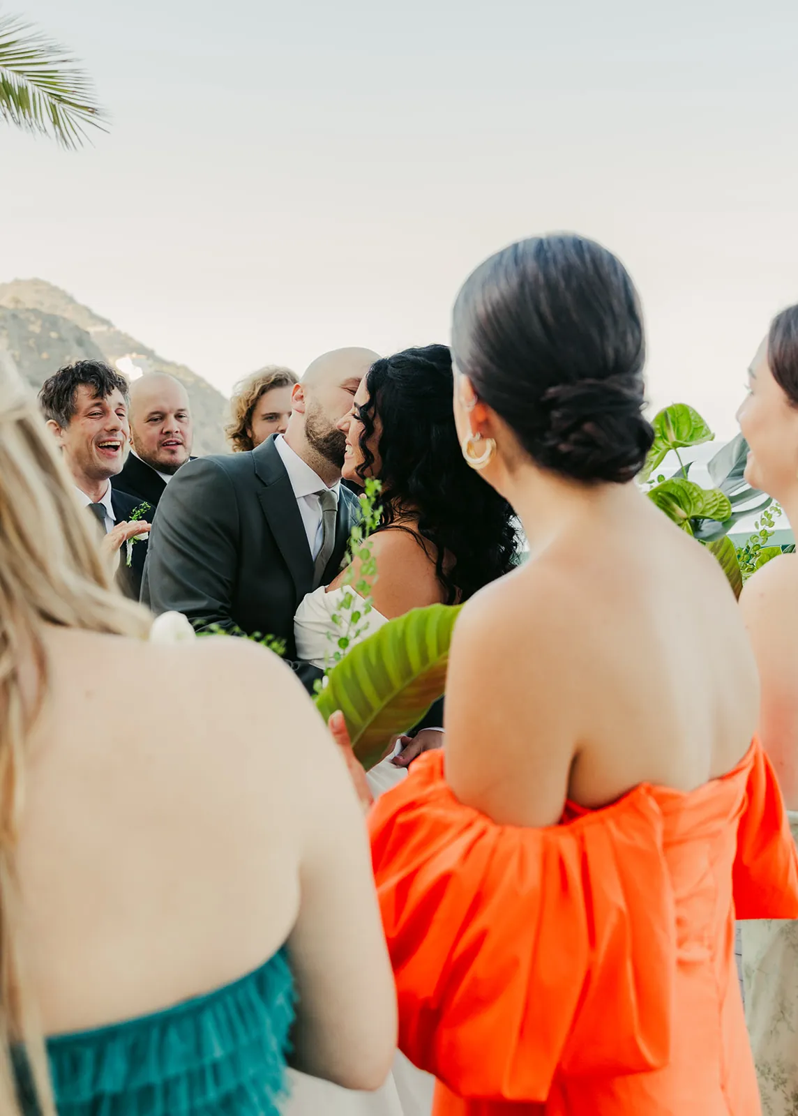 Candid moment of bride and groom kissing surrounded by their laughing bridal party at their Catalina Island destination wedding, Catalina hills visible behind them
