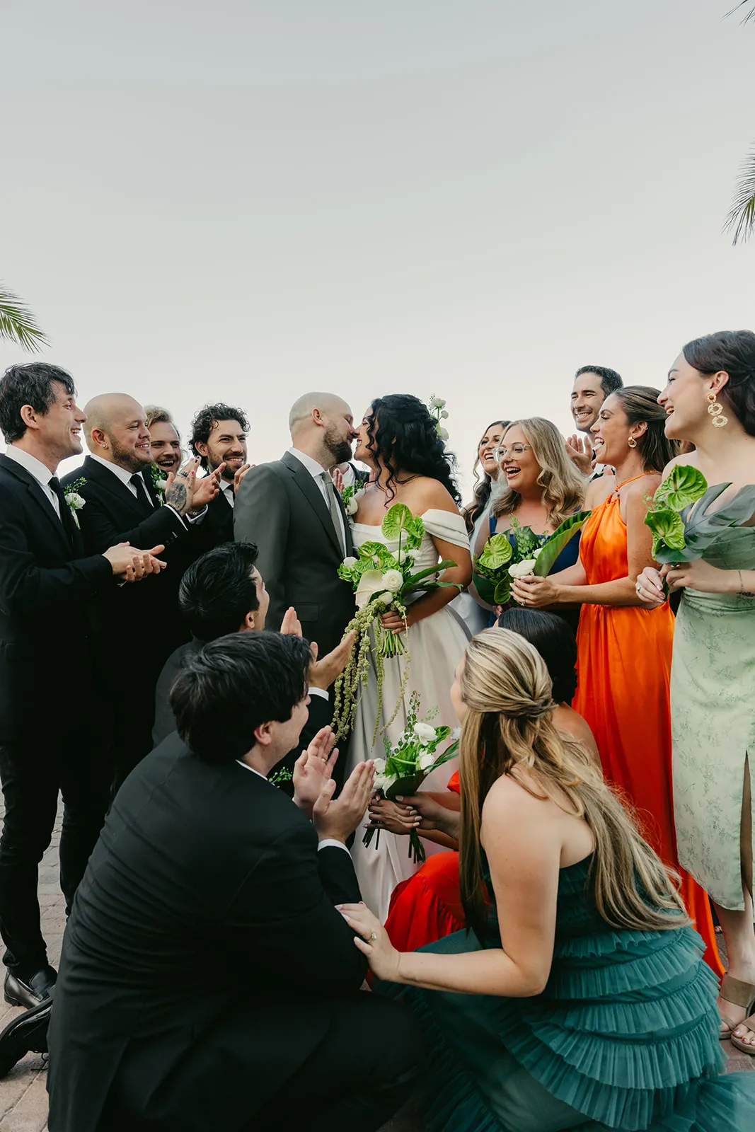 Full bridal party surrounding the bride and groom as they share a kiss outside the Catalina Island Casino, colorful gowns and tropical anthurium bouquets, Catalina hills in the background