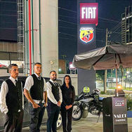 Quatro funcionários uniformizados (três homens e uma mulher) do Circuit Park Valet, vestindo coletes pretos, estão em pé ao lado de um totem de estacionamento e uma motocicleta, sob um guarda-sol à noite. Ao fundo, um grande letreiro iluminado das marcas Fiat e Abarth, com as cores da bandeira italiana em uma coluna.