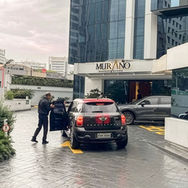 Um funcionário de uniforme escuro abre a porta de um Mini Cooper Countryman preto na entrada do edifício 'Murano Business Office'. Um sinal do 'Circuit Park Estacionamento' com informações de preço está visível à esquerda, e o chão está molhado, sugerindo que choveu.