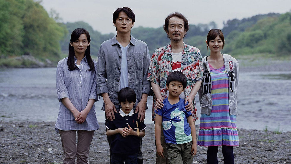 A group of six people, including two children, stand by a river. They're dressed casually, with a backdrop of trees and water on a cloudy day.