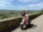 A smiling couple poses with a vespa in tuscany overlooking lush green hills and a blue sky, exuding a joyful and scenic vibe.