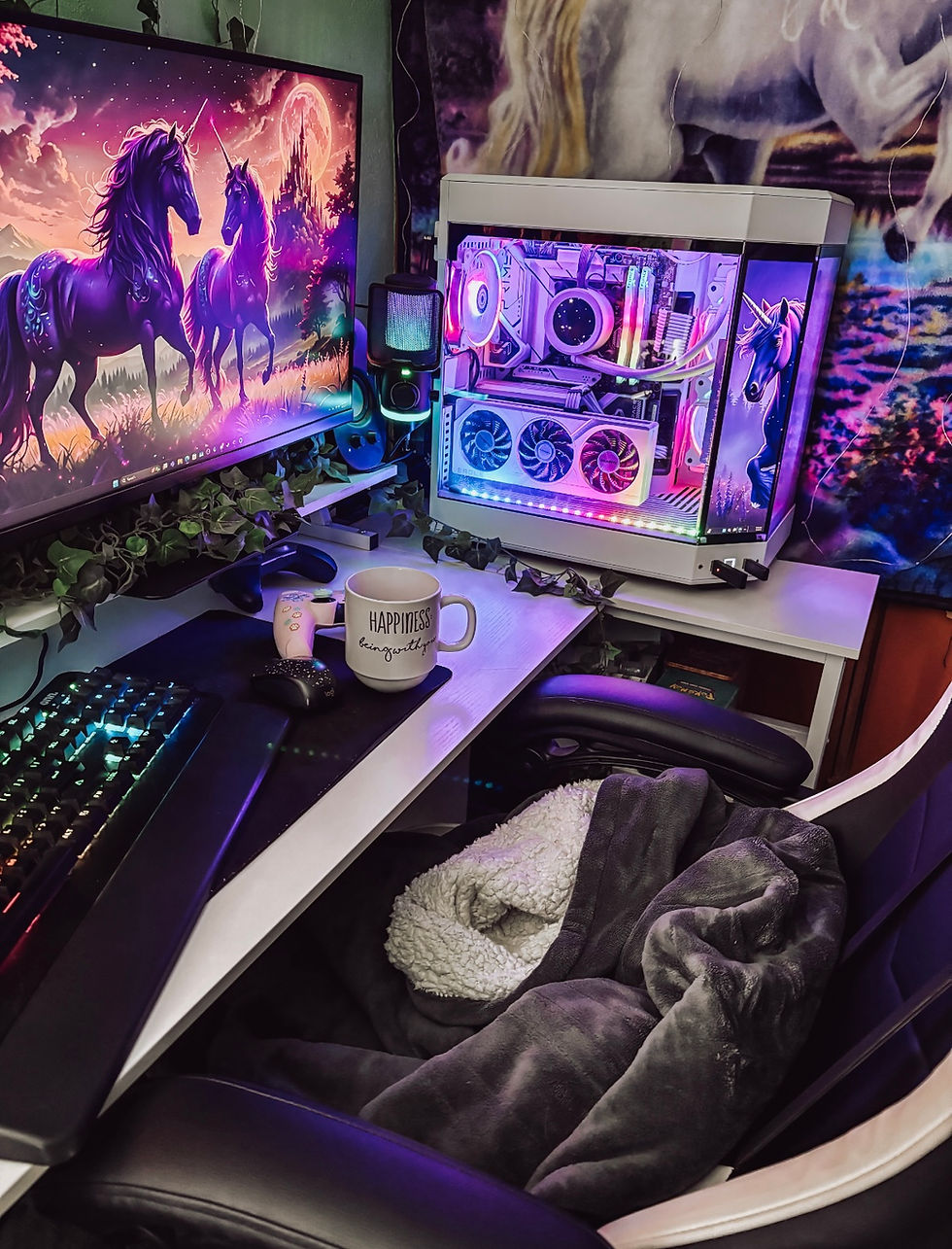 Cozy pastel gaming setup featuring a dual-monitor display with unicorn wallpaper, RGB lit PC, and chair draped with a soft heated blanket.