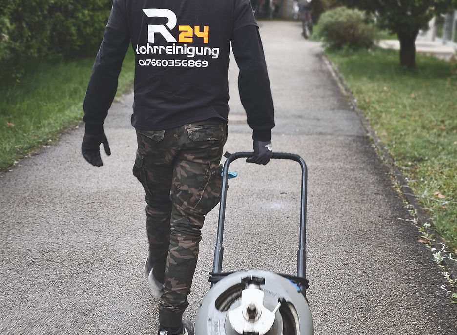 Ein Mitarbeiter von R24 Rohrreinigung läuft einen gepflasterten Weg entlang und zieht eine motorisierte Rohrreinigungsmaschine auf Rädern hinter sich her. Er trägt eine schwarze Kapuzenjacke mit dem Logo und der Telefonnummer von 'R24 Rohrreinigung' auf dem Rücken sowie eine Camouflage-Hose und schwarze Handschuhe. Der Weg ist von grünen Hecken und Büschen gesäumt, daneben stehen mehrstöckige Wohnhäuser. Das Wetter ist bewölkt.