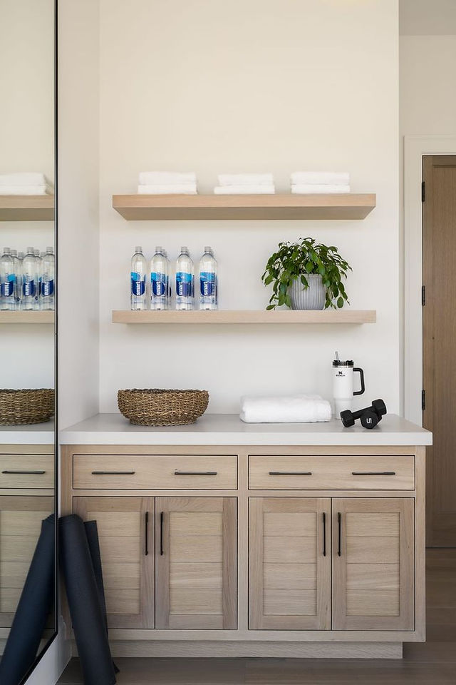 Wood built-in cabinets in home gym with wall shelving, bottled waters, and towels