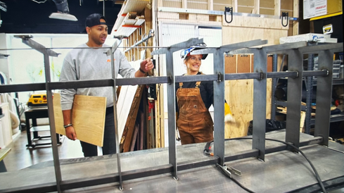 MAn and woman look at metal welded table base in workshop setting.