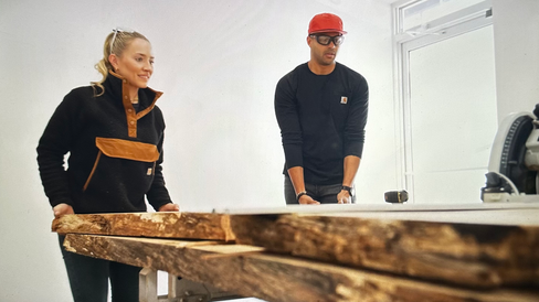 Man and woman look over large wood slab in white room.