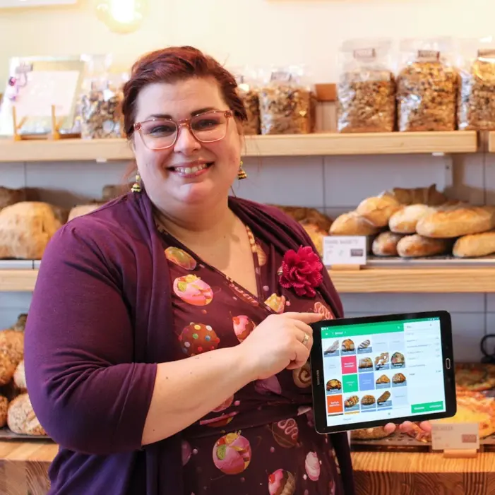 Femeie zâmbitoare într-o brutărie, prezentând o tabletă cu software POS.