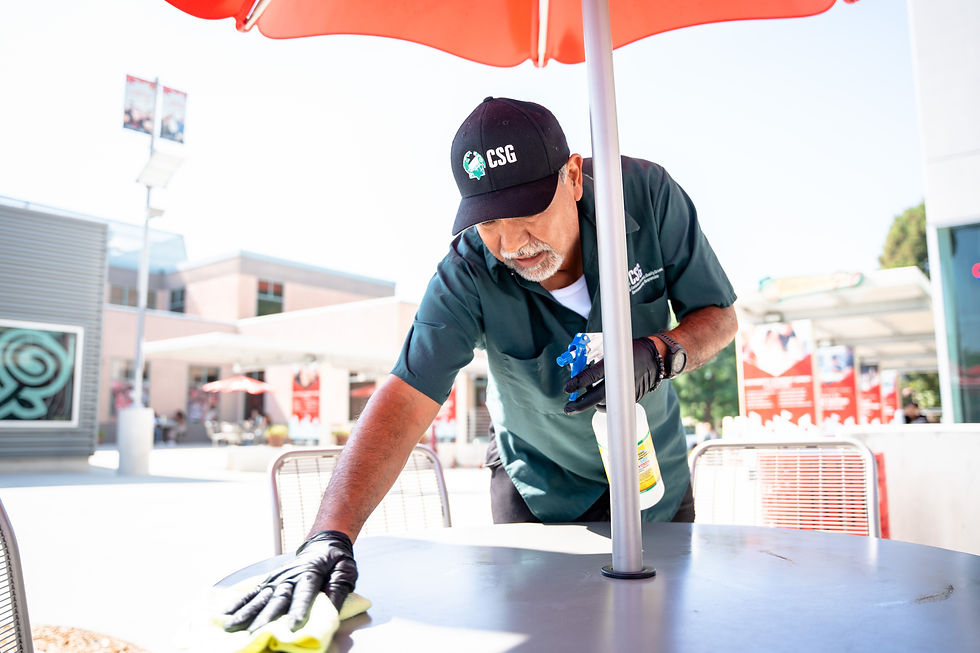 A person in a green uniform and black gloves cleans an outdoor table with a spray bottle and cloth under a red umbrella.
