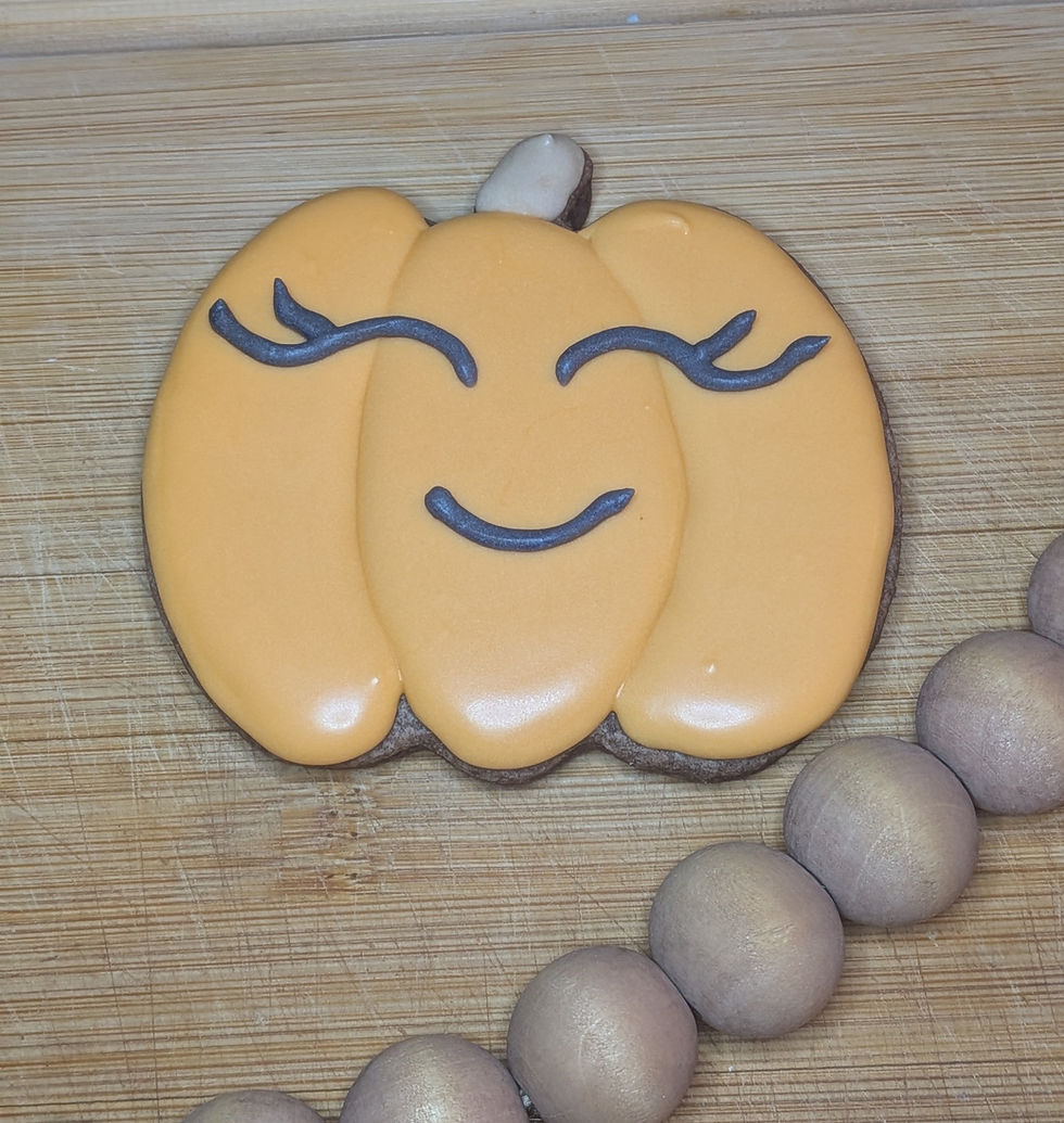 Smiling pumpkin-shaped cookie with orange dye-free icing and black features on a wooden board, next to a string of round, brown wooden beads.