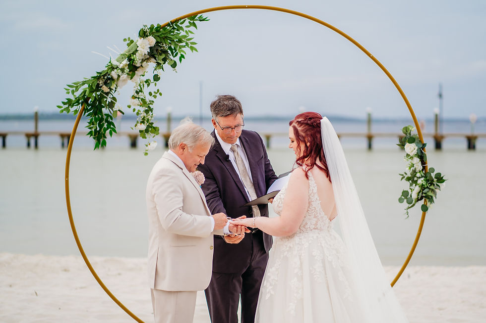 A couple exchanges rings on a beach under a floral arch, overlooking the water. An officiant guides the ceremony. The mood is joyful.
