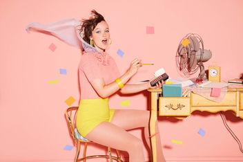 A woman is sitting at a desk in front of a fan showcasing hair and makeup artistry by Piper von Hoene. 