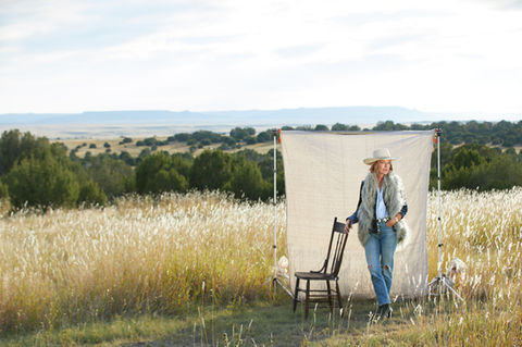 Woman in a white cowboy hat and fur vest standing in a field with a chair and a backdrop behind her.