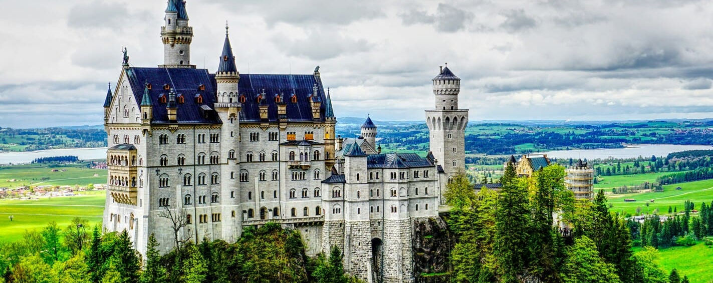 Білосніжний замок Нойшванштайн на скелястому пагорбі, оточений густим зеленим хвойним лісом та легким туманом.