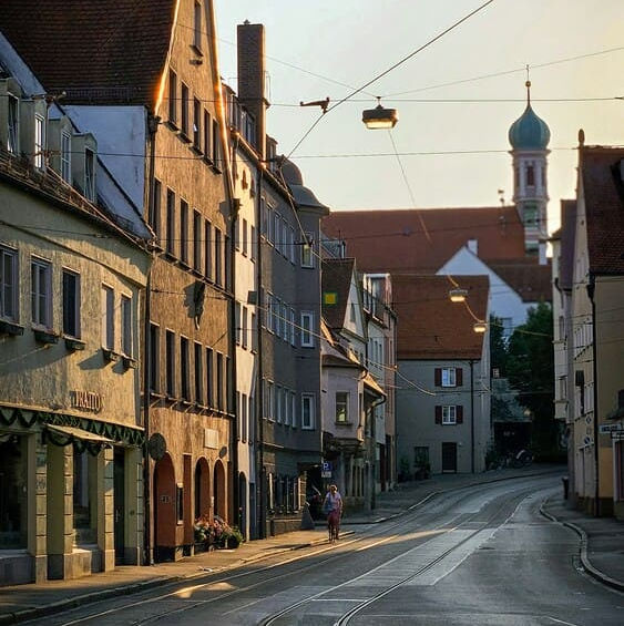 Звивиста вулиця з трамвайними коліями та старовинними будинками в Аугсбурзі, залита теплим вечірнім сонячним світлом.