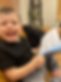 A smiling toddler boy holding a Unique I Am book while sitting on a couch with his cute chubby toes touching.