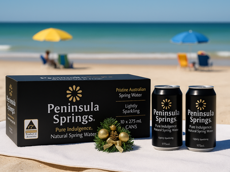 Peninsula Springs lightly sparkling spring water cans and box displayed on a sandy Australian beach with festive Christmas decorations, ocean waves, and colourful umbrellas in the background