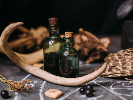 Two dark glass bottles with corks sit inside a large antler. Feathers, black beads, and dried herbs on a gray surface create an earthy vibe.