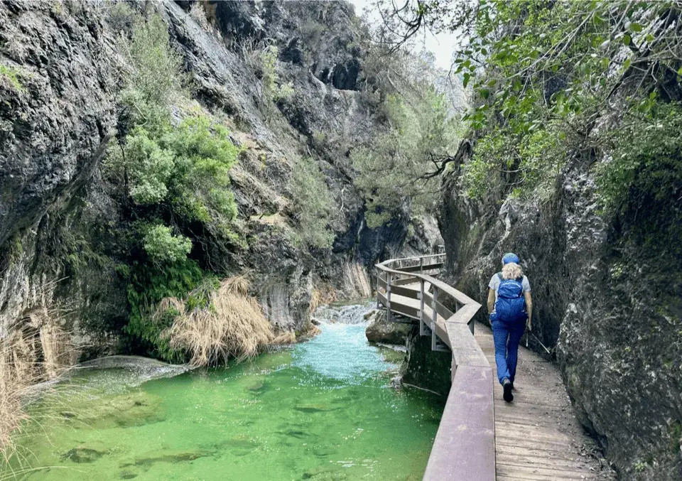 Persona caminando por pasarela de madera junto a río de aguas turquesas en un cañón rocoso.
