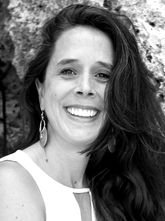 A smiling white woman with side-parted long dark hair looks into the camera. She's wearing a sleeveless white top, and in the background are textured rocks.