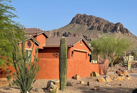 Southwestern-style home in Tucson Estates, Arizona, surrounded by desert landscaping with tall saguaros, gravel paths, and scenic views of rugged mountains in the distance. The warm-toned house and native plants reflect the area’s blend of residential charm and natural Sonoran beauty.