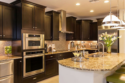 A Tucson kitchen featuring dark wood cabinets, granite countertops, and stainless steel appliances, including a double oven and side-by-side refrigerator. Pendant lights hang over a central island adorned with fresh flowers, while small appliances and glassware add lived-in warmth to the clean, well-organized space.