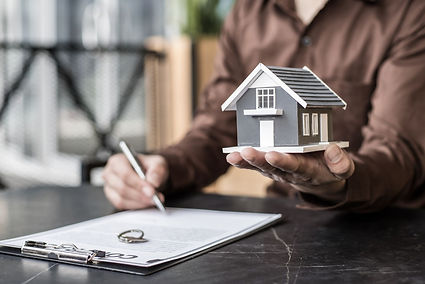 A person signs a contract while holding a small model home in their other hand, framed by warm lighting and a clean workspace. The scene reflects a property transaction in Tucson, blending professionalism and care—ideal for communicating trust in estate services or home transition support.