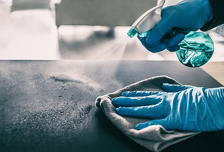 A person wearing blue gloves disinfects a bathroom counter in a Tucson home, spraying cleaning solution and wiping down a bright, polished surface. Brass fixtures and pendant lights frame the moment, highlighting attention to detail and the quiet impact of a freshly cleaned space.