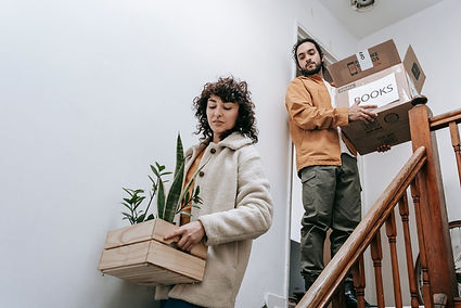 Two people carry boxes and plants down a staircase in a Tucson home, signaling a tenant move-out in progress. The wooden steps and indoor lighting frame the transition as the space is cleared for its next chapter.