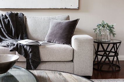A Tucson living room with a light gray sofa draped in a dark gray throw and styled with a studded velvet pillow, paired with a round black side table holding a clear vase of greenery. The space is softly lit and grounded by warm wood tones and ceramic decor, creating a clean, welcoming atmosphere that balances comfort with quiet sophistication.