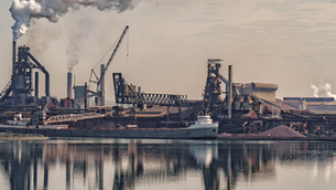 Usine sidérurgique au bord de l’eau, cargo à quai, cheminées fumantes et reflets calmes sur l’eau.