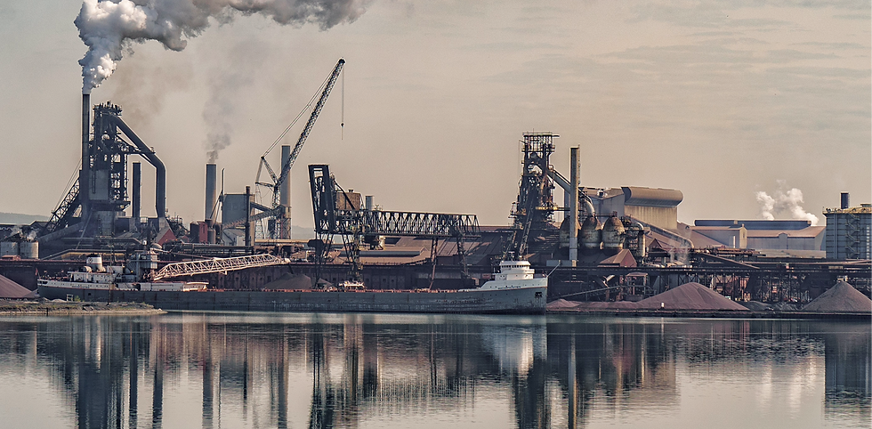 Usine sidérurgique au bord de l’eau, cargo à quai, cheminées fumantes et reflets calmes sur l’eau.
