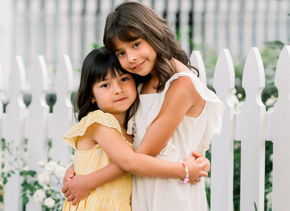Sisters hug in front of white picket fence.
