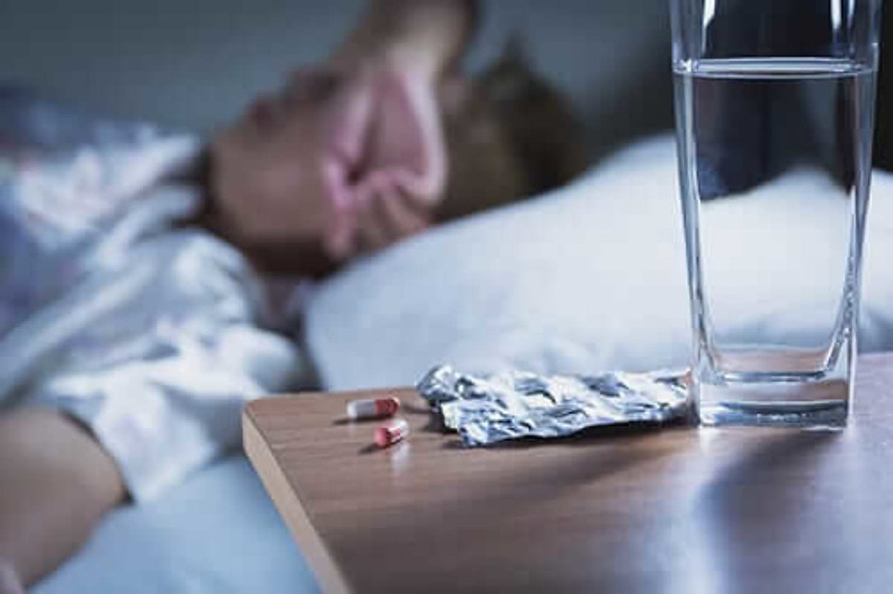 Pessoa aparenta estar preocupada e indisposta em sua cama, ao lado e em consequência dos remédios que tomou para dormir.