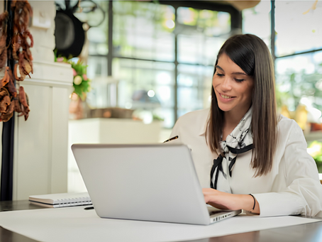 student working on a laptop in a bright, modern office. Notebook and pen on the desk, green plants in the background.