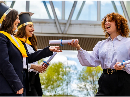 Graduates in caps and yellow sashes receive diplomas outdoors, smiling under a glass bridge, with a bright, cloudy sky and trees behind.