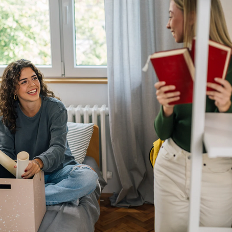 Two students in room unpacking. One sits on bed with rolled paper, smiling at standing girl holding red books. Bright window light.