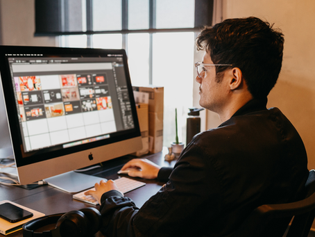an engineer with glasses edits a design layout on an iMac in a warm office, focused at his desk with keyboard and mouse.