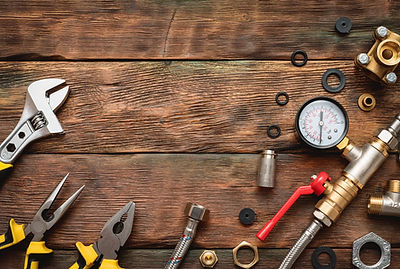 An overhead photo of various plumbing tools and components arranged around the perimeter of a rustic wooden plank surface, leaving a blank space in the center. The items include a chrome adjustable wrench, pliers, assorted gaskets and O-rings, brass and chrome pipe fittings, a pressure gauge with a red needle, and a silver hose with a red-handled valve.