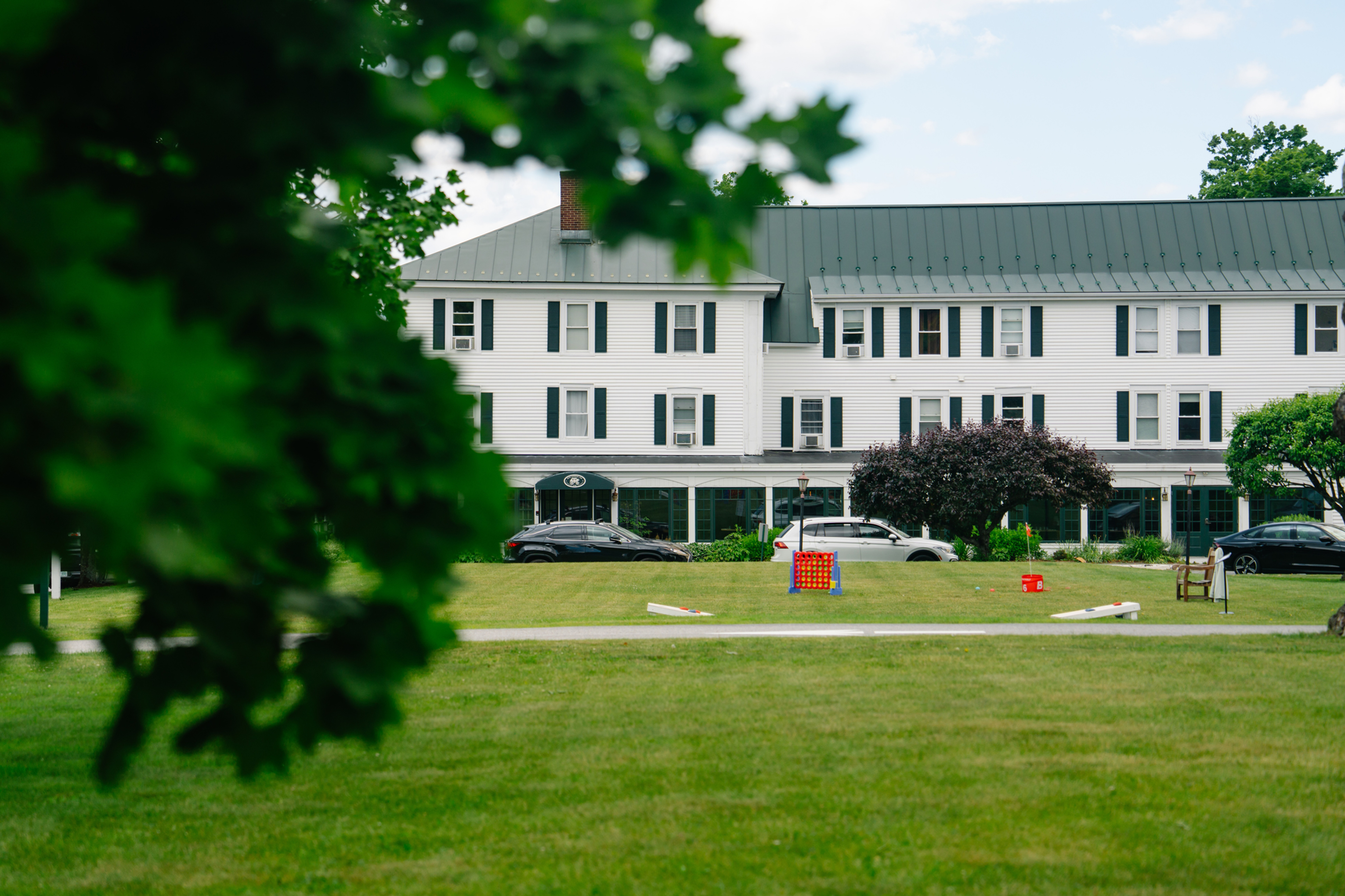 New London Inn's white clapboard building with dark green shutters and metal roof, seen across a lawn with lawn games including giant Connect Four and cornhole, framed by tree branches in the foreground.