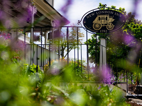 The exterior sign of New London Inn photographed through a soft blur of purple spring wildflowers and lush green foliage in the foreground. The oval black iron sign reads "New London Inn" with an illustration of a horse-drawn stagecoach and the founding year 1792. Behind the sign, a white clapboard porch with hanging flower baskets and black iron railings is visible beneath a bright blue sky and green trees.
