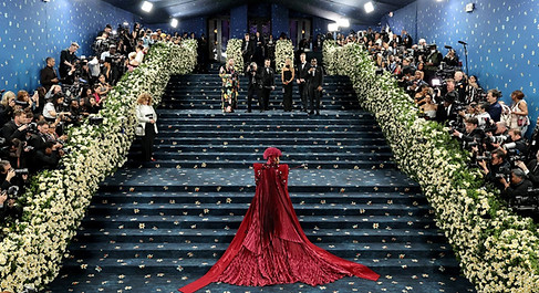 Woman in a dramatic red gown standing on a monumental staircase surrounded by formally dressed guests.