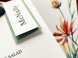 Close-up of a name card with "McNally" in green text attached to a menu. Background features a colorful floral design.
