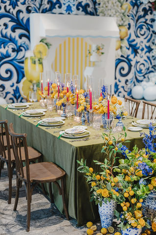 Mediterranean-inspired dining table with lemons, blue and yellow florals, ceramic vases, taper candles, and green linen tablecloth set against a tile-patterned backdrop.
