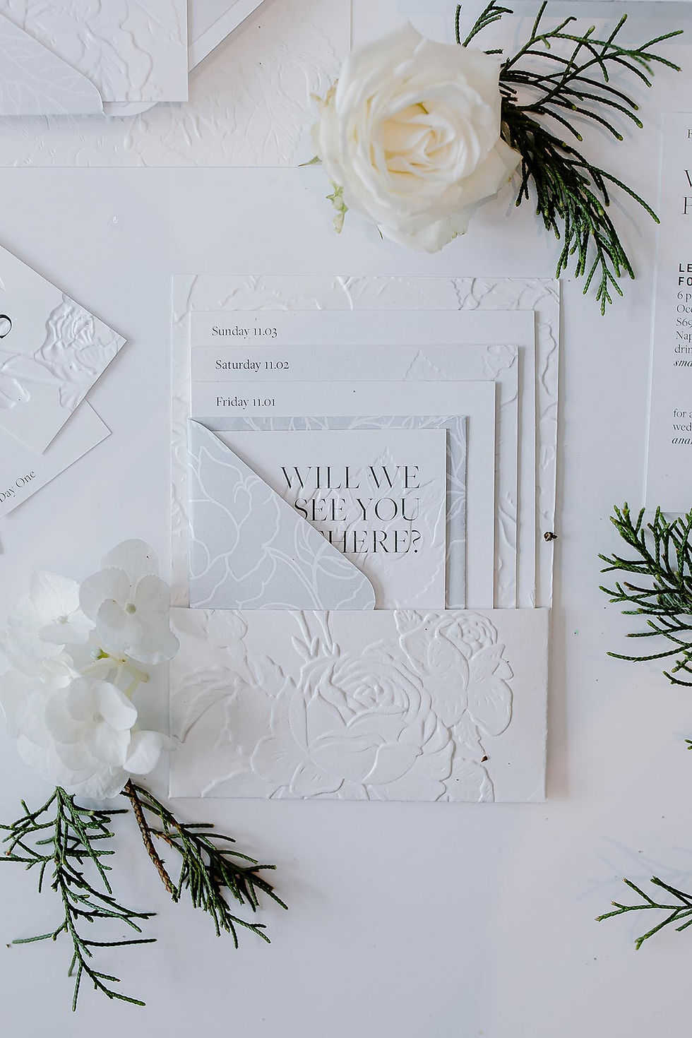 White embossed wedding weekend stationery with layered schedule cards, floral paper details, and a “Will We See You There?” insert