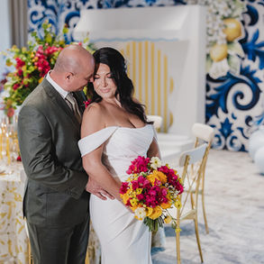 Bride and groom embracing in front of Mediterranean-themed reception setup with bright floral bouquet.
