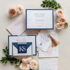 Blue and white wedding invitation suite with embossed floral details, vellum wrap, monogram piece, tassel tag, and blush roses arranged on a neutral background
