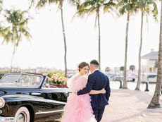 Couple embraces near a classic black car, with palm trees in the background. The woman wears a pink gown, creating a romantic vibe.
